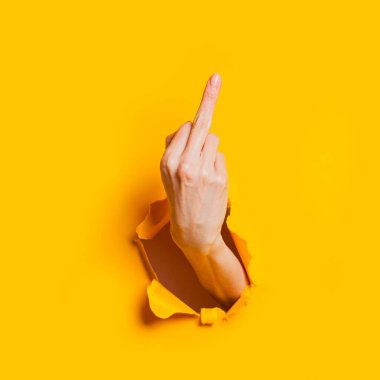 Female hand showing gesture meaning fuck you or fuck off through the hole in the yellow paper background.