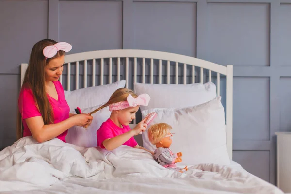 Женщина заплетает косички дочери, сидящей на кровати — стоковое фото