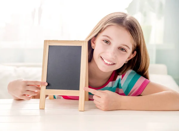 Boş küçük kara tahta ile masadan gülümseyen küçük kız — Stok fotoğraf
