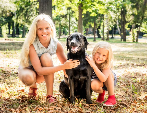 Les petites filles blondes s'assoient serrant un chien noir dans un parc — Photo