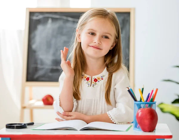 Маленькая блондинка поднимает руку на ответ, сидя за столом в школьном классе — стоковое фото