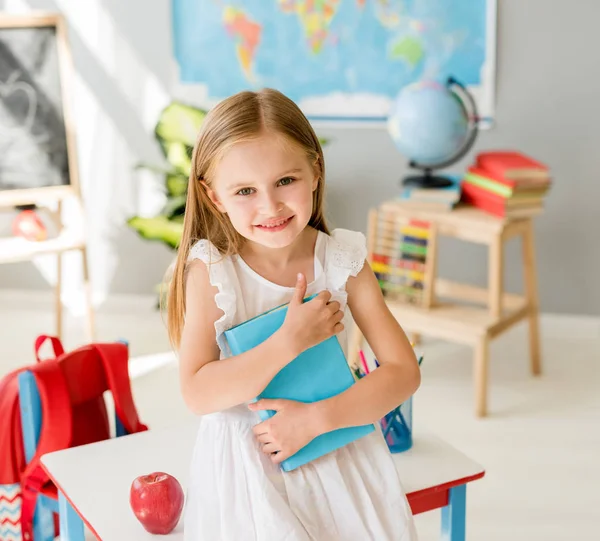 Usměvavá blonďatá holčička drží blue book ve školní třídě — Stock fotografie