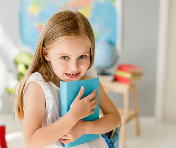 Pequena menina loira sorridente segurando livro azul na classe da escola — Fotografia de Stock