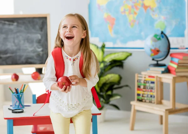 Pequeña rubia riendo sosteniendo una manzana en el aula de la escuela — Foto de Stock