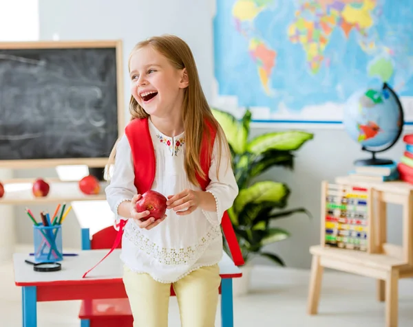 Kleines lachendes blondes Mädchen mit einem Apfel im Klassenzimmer — Stockfoto