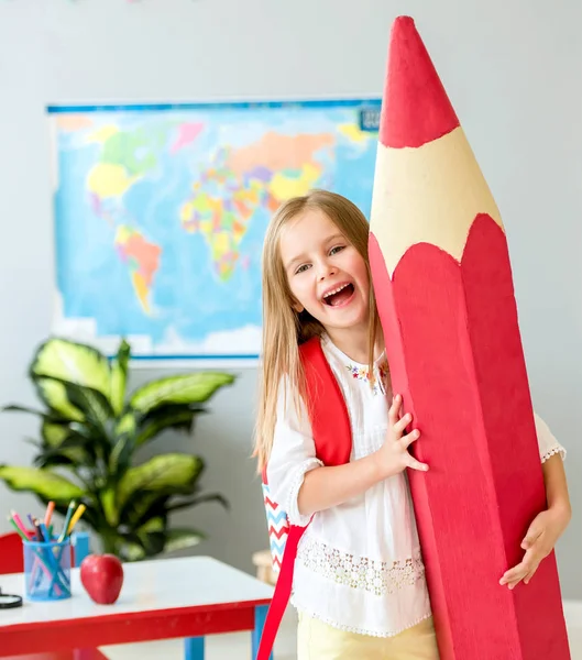 Pouco sorrindo menina loira segurando enorme lápis vermelho na sala de aula da escola — Fotografia de Stock