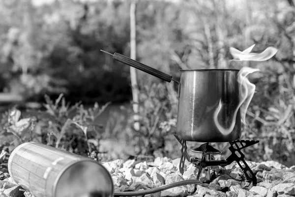 Portable tourist gas stove with a gray kettle on a background of nature in the mountains. Camping kitchen and tea. Unsafe gas stpve. Black and white image.