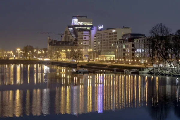 Riga, Letônia - 29 de abril de 2018: Cidade da Noite no Banco do Rio Daugava. Urban View Of National Library Building and Radisson Blue Daugava (em inglês). Foto noturna . — Fotografia de Stock