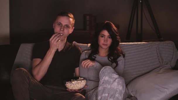 Молодая пара смотрит что-то смешное по телевизору дома и ест попкорн — стоковое видео
