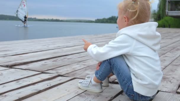 Menina Bonita Mostrando Windsurfista Com Dedo Enquanto Sentada Perto Lago — Vídeo de Stock