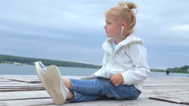 漂亮的小女孩看着平板电脑和湖 而坐在木码头慢动作 — 图库视频影像