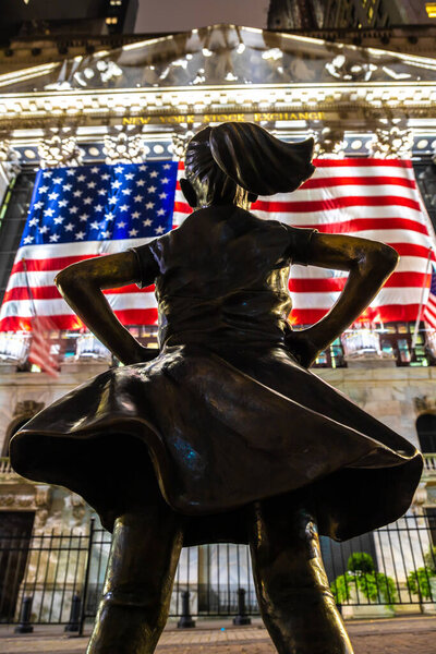 NEW YORK CITY, USA - MARCH 15, 2020:  Fearless Girl Statue looking up at  New York Stock Exchange building at Wall Street in Manhattan at night, New York City, USA