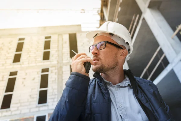 男性总工程师在检查施工现场时 使用对讲机佩戴白色安全安全帽 发展和建筑业理念 免版税图库照片