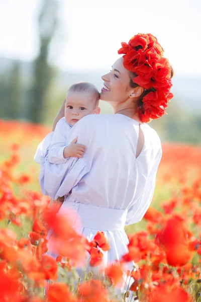 彼女の腕の中で小さな息子と白ロングドレスと新鮮な赤いケシの花輪の美しい若い母親は 夏の日に 日当たりの良い巨大な牧草地を歩く開花赤花の美しさを楽しむ — ストック写真