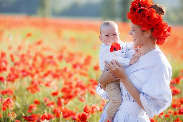 彼女の腕の中で小さな息子と白ロングドレスと新鮮な赤いケシの花輪の美しい若い母親は 夏の日に 日当たりの良い巨大な牧草地を歩く開花赤花の美しさを楽しむ — ストック写真
