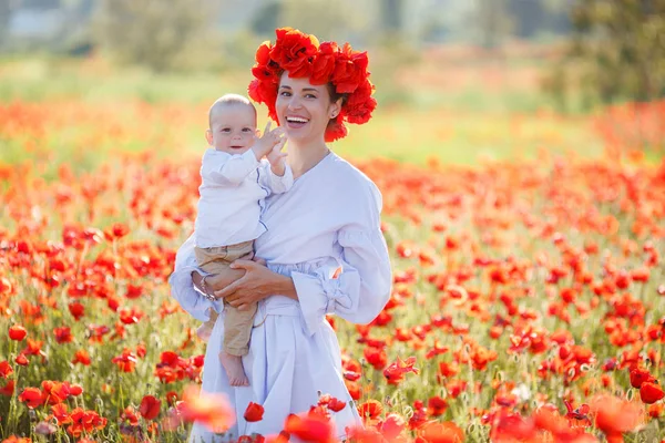 彼女の腕の中で小さな息子と白ロングドレスと新鮮な赤いケシの花輪の美しい若い母親は 夏の日に 日当たりの良い巨大な牧草地を歩く開花赤花の美しさを楽しむ — ストック写真