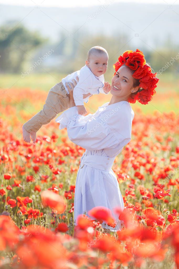 A beautiful young mother in a white long dress and a wreath of fresh red poppies with a small son in her arms enjoy the beauty of flowering red flowers, walking through a huge meadow on a sunny, summer day