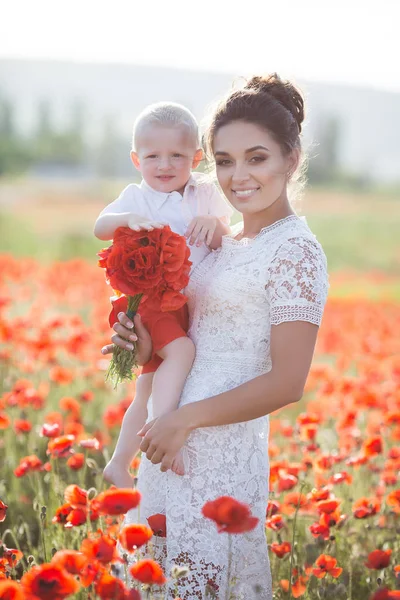 彼女の腕で若い息子と白ドレスの美しい若い母親は 日当たりの良い夏の日に巨大な牧草地を歩く赤い咲きの美花を楽しんでいます 幸せな母と赤いケシのフィールドで息子の夏のシーン — ストック写真