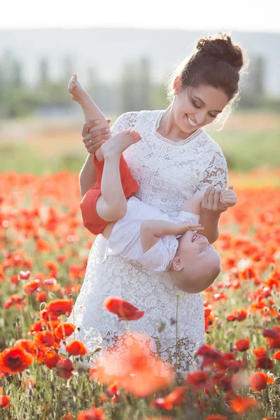 彼女の腕で若い息子と白ドレスの美しい若い母親は 日当たりの良い夏の日に巨大な牧草地を歩く赤い咲きの美花を楽しんでいます 幸せな母と赤いケシのフィールドで息子の夏のシーン — ストック写真