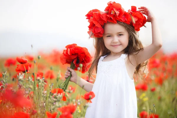 長い巻き毛と白のノースリーブ ドレスを着て 茶色の目を持つ幼児女の子は赤咲くポピーと大規模なフィールドに田舎で単独で彼女の時間を費やしている彼女の手でケシの花の花束を持って身に着けている赤い花の花輪 — ストック写真