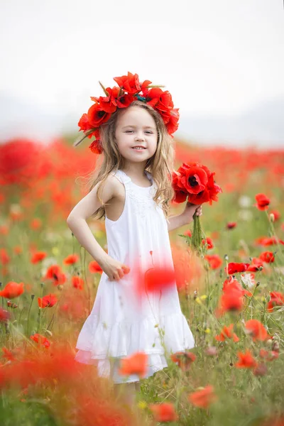 長い巻き毛と白のノースリーブ ドレスを着て 茶色の目を持つ幼児女の子は赤咲くポピーと大規模なフィールドに田舎で単独で彼女の時間を費やしている彼女の手でケシの花の花束を持って身に着けている赤い花の花輪 — ストック写真