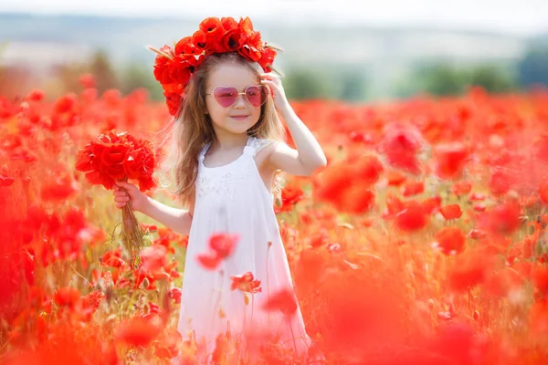 長い巻き毛 白いノースリーブのドレスに身を包んだ小さな幼児女の子ピンクのメガネを身に着けている 赤いポピーで大規模なフィールドの田舎で一人の時間を費やしている 赤い花の花輪を身に着けている 彼女の手でケシの花の花束を保持 — ストック写真