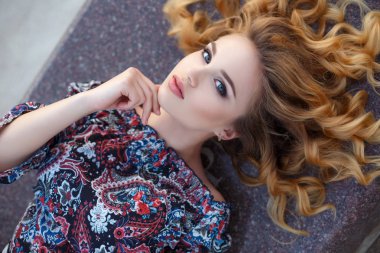 Uzun kıvırcık saçlı, Sarışın, uzun siyah kirpik ve hafif makyaj, şirin gülümseme, çok renkli desenleri ile karanlık bir bluz giymiş güzel genç kadın açık omuz, açık havada yaz saati bir şehir sokak üzerinde poz