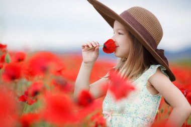 Küçük beyaz hafif kolsuz elbise kız, sarı saçlı, kafasında büyük ağzına kadar bir poz, ayakta açık havada yaz aylarında çiçek açan büyük bir alan üzerinde dağlık bir alanda kırmızı haşhaş ile kahverengi güneş şapkası giyiyor