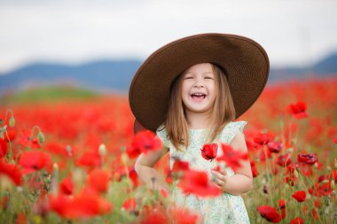 Küçük beyaz hafif kolsuz elbise kız, sarı saçlı, kafasında büyük ağzına kadar bir poz, ayakta açık havada yaz aylarında çiçek açan büyük bir alan üzerinde dağlık bir alanda kırmızı haşhaş ile kahverengi güneş şapkası giyiyor