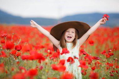 Küçük beyaz hafif kolsuz elbise kız, sarı saçlı, kafasında büyük ağzına kadar bir poz, ayakta açık havada yaz aylarında çiçek açan büyük bir alan üzerinde dağlık bir alanda kırmızı haşhaş ile kahverengi güneş şapkası giyiyor