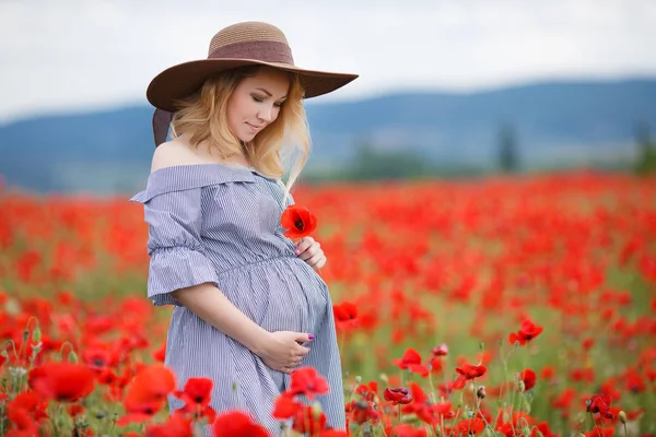 青い縞の白いドレスに身を包んだ大きな腹 ブロンドのかわいい妊婦 夏に屋外でポーズをする大きな茶色の太陽の帽子をかぶった彼の頭の上に 花のポピーの紅海の中で1つの山の野原に立っている — ストック写真