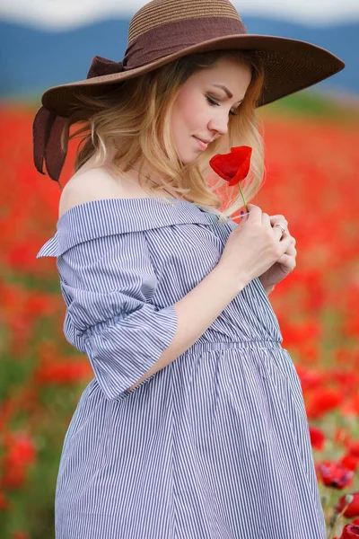 青い縞の白いドレスに身を包んだ大きな腹 ブロンドのかわいい妊婦 夏に屋外でポーズをする大きな茶色の太陽の帽子をかぶった彼の頭の上に 花のポピーの紅海の中で1つの山の野原に立っている — ストック写真
