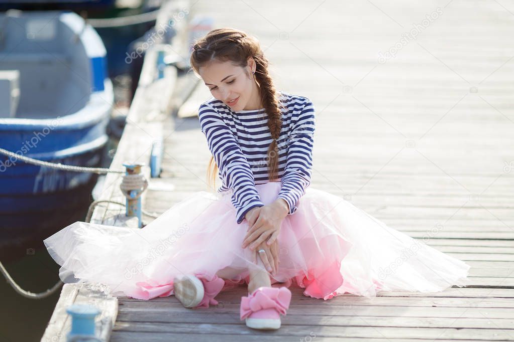 A beautiful young woman, a brunette with long hair plaited in a braid, dressed in a pink transparent skirt and a striped moth jersey, spends time sitting on a wooden pier by the sea next to the moored boat