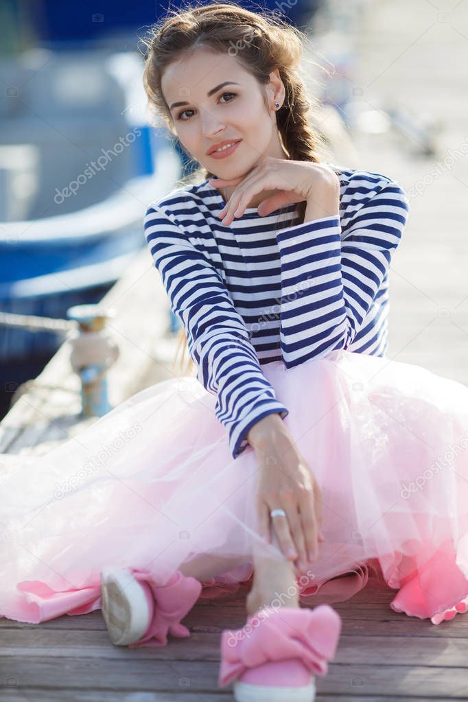 A beautiful young woman, a brunette with long hair plaited in a braid, dressed in a pink transparent skirt and a striped moth jersey, spends time sitting on a wooden pier by the sea next to the moored boat