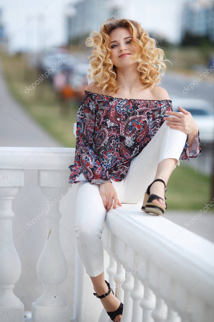 Beautiful young woman with long curly hair, blond, long black eyelashes and light make-up, cute smile, dressed in a dark blouse with multi-colored patterns, open shoulders, posing outdoors in the summer time on a city street