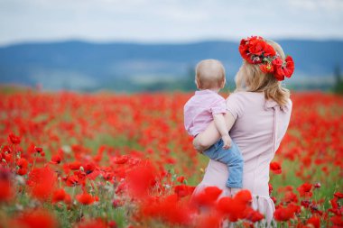 Mutlu genç hamile kadın, Sarışın, bir çelenk taze çiçek haşhaş açık havada dağlarda vahşi kırmızı poppies, oğlunun elinde büyük bir alan üzerinde zaman harcıyor onun baş giyer pembe elbise giymiş