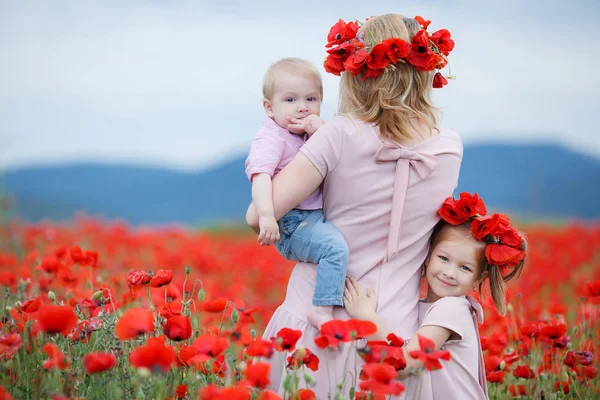 幸せな 妊娠中の女性がピンクのドレスに身を包んだ新鮮なポピーの花の花輪を身に着けている屋外巨大な赤い花の花輪と小さな息子手の小さな娘と野生の赤いケシ分野に時間を費やして 彼女の頭の上 — ストック写真