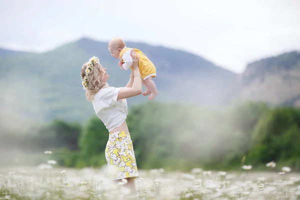 A happy mother holds in her arms her newborn son standing outdoors in the highlands among a blooming chamomile field. Family, mother and baby, against the backdrop of mountains and blue sky on a white meadow of blooming daisies