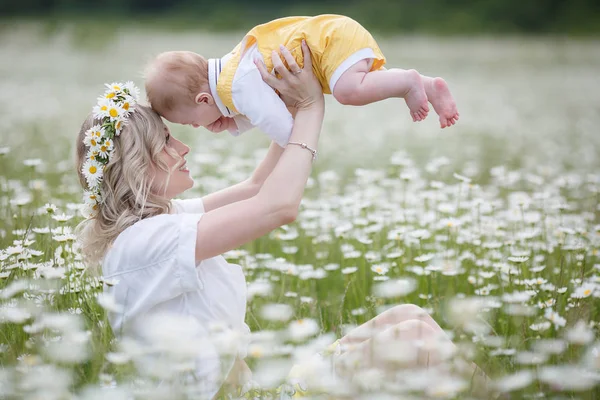 一位快乐的母亲抱着刚出生的儿子 站在户外的高地里 在一片盛开的洋甘菊地里 母亲和婴儿 在山和蓝天的背景下 在盛开的雏菊的白色草地上 — 图库照片