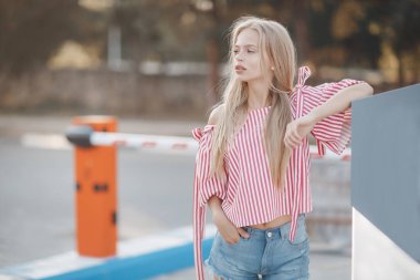 Uzun, güzel bir genç kadınla pink düz saç ve gri gözler, güzel makyaj, dolgun dudaklar, tatlı bir gülümseme, Sarışın, ona vakit şehirde, yaz aylarında açık havada beyaz mayoyu ve güneş ışığında poz kareli gömlek giymiş
