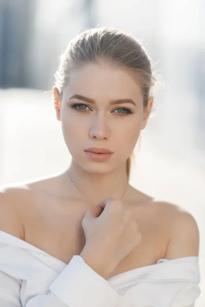 A young beautiful woman with long, hair and gray eyes, beautiful makeup, pink plump lips, sweet smile,blonde hair, spending her time outdoors in the summer in the city, dressed in a white blouse, posing in the sunlight. Portrait of happy young woman