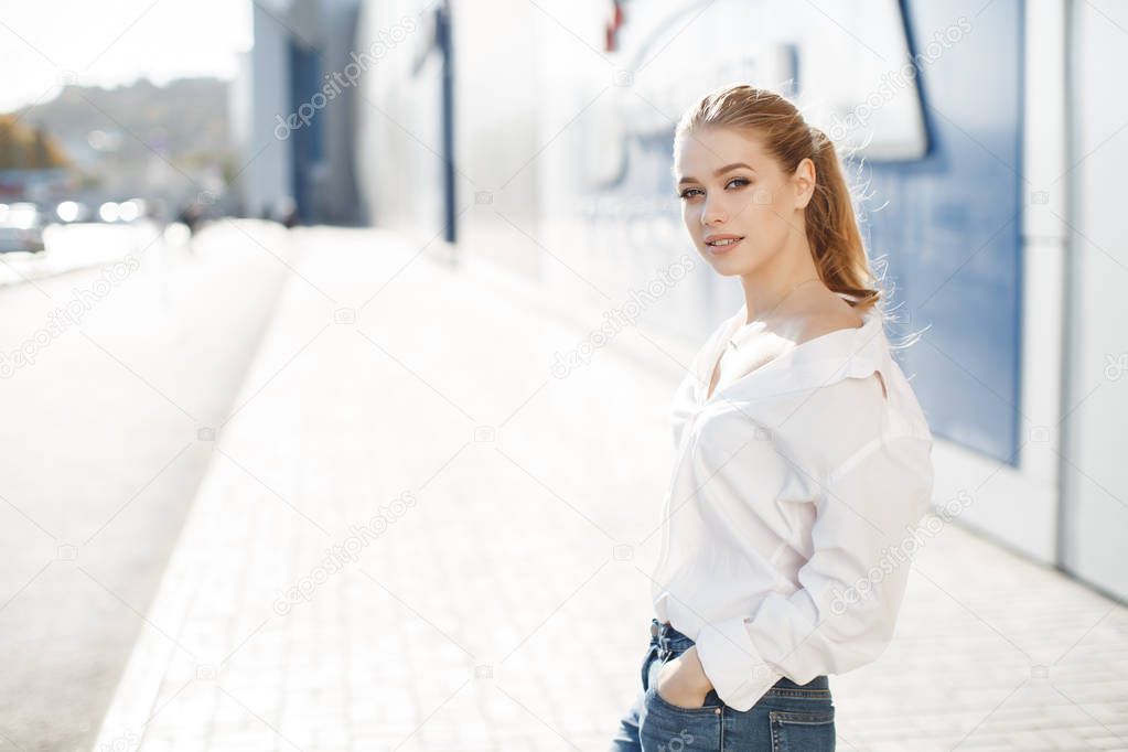 A young beautiful woman with long, hair and gray eyes, beautiful makeup, pink plump lips, sweet smile,blonde hair, spending her time outdoors in the summer in the city, dressed in a white blouse, posing in the sunlight. Portrait of happy young woman