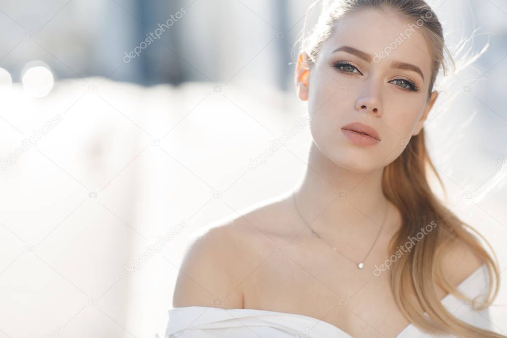 A young beautiful woman with long, hair and gray eyes, beautiful makeup, pink plump lips, sweet smile,blonde hair, spending her time outdoors in the summer in the city, dressed in a white blouse, posing in the sunlight. Portrait of happy young woman