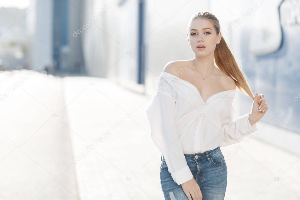 A young beautiful woman with long, hair and gray eyes, beautiful makeup, pink plump lips, sweet smile,blonde hair, spending her time outdoors in the summer in the city, dressed in a white blouse, posing in the sunlight. Portrait of happy young woman