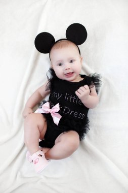 Şirin bebek girllying yalnız beyaz bir yatağa giyinmiş. Küçük çocuk kameraya bakıyor. Beyaz arka plan üzerinde kepçe öğrenen bebek. Mutlu kız portresi. Bebek moda giysileri. 5 aylık bir çocuk tek başına, bembeyaz bir yatakta yatarken eğleniyor.