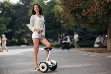 Güzel kadın güneş gözlüğü, beyaz bir bluz ve şort zaman şehir parkı açık havada yaz aylarında bir segway sürme harcıyor. Yeşil Park Elektrik mini vurgulu kurulu scooter ile genç esmer kız. İyi bir hava trendy ulaşım teknolojisinin