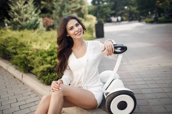 Beautiful woman in sunglasses, in a white blouse and shorts spends time in the city park outdoors in the summer,riding a segway.Young brunette girl with electrical mini hover board scooter in green park. Good  weather trendy transportation technology