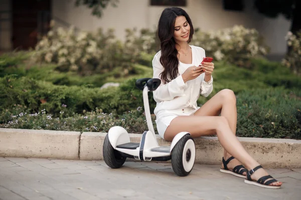 Beautiful woman in sunglasses, in a white blouse and shorts spends time in the city park outdoors in the summer,riding a segway.Young brunette girl with electrical mini hover board scooter in green park. Good  weather trendy transportation technology