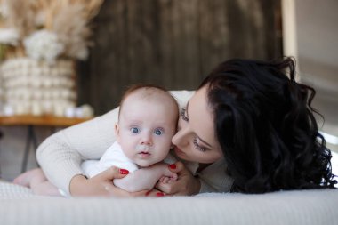 Yeni doğmuş çocuğunu tutan genç anne. Kadın ve yeni doğan çocuk beyaz bir yatak odasında rahatlayın. Genç bir anne kucağında yeni doğan oğlunu nazikçe tutar. Evde aile. Yeni doğan oğlunu whith oynayan genç anne