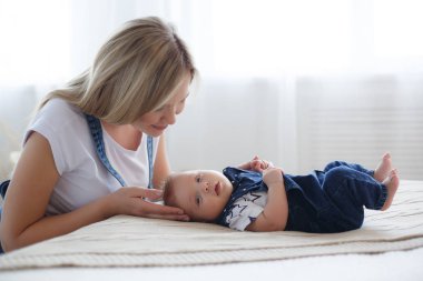 Yeni doğmuş çocuğunu tutan genç anne. Kadın ve yeni doğan çocuk beyaz bir yatak odasında rahatlayın. Genç bir anne kucağında yeni doğan oğlunu nazikçe tutar. Evde aile. Yeni doğan oğlunu whith oynayan genç anne